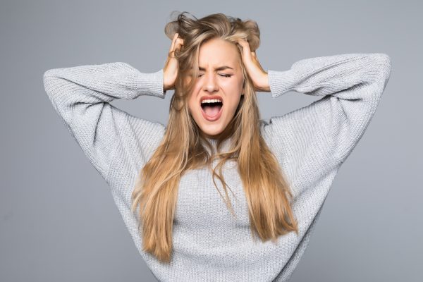 Portrait of a frustrated angry woman screaming out loud and pulling her hair out isolated on the gray